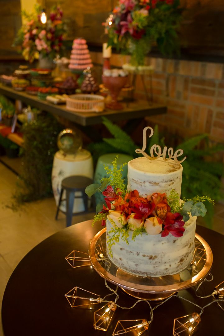 White wedding cake topped with 'Love' and fresh flowers on display with string lights and dessert table in background.