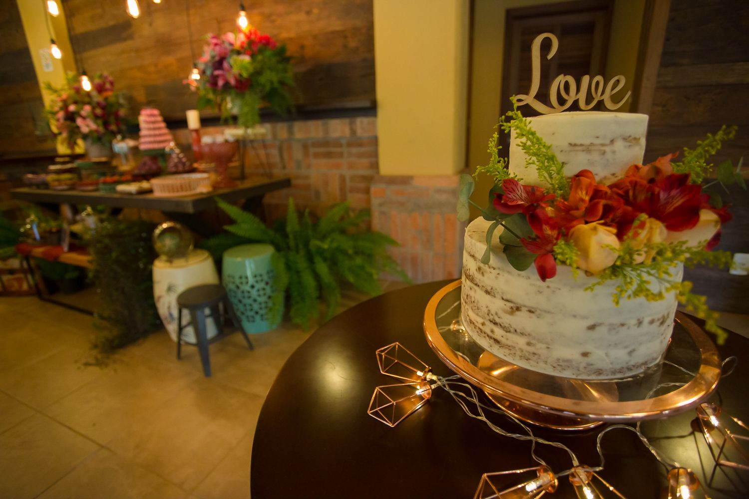 Rustic wedding cake display with geometric lights and 'Love' topper against dessert table backdrop.