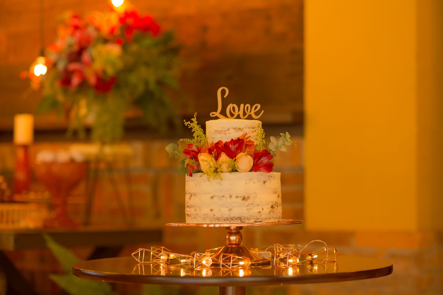 Wedding cake with 'Love' topper and fresh flowers illuminated by warm lighting on dark table.