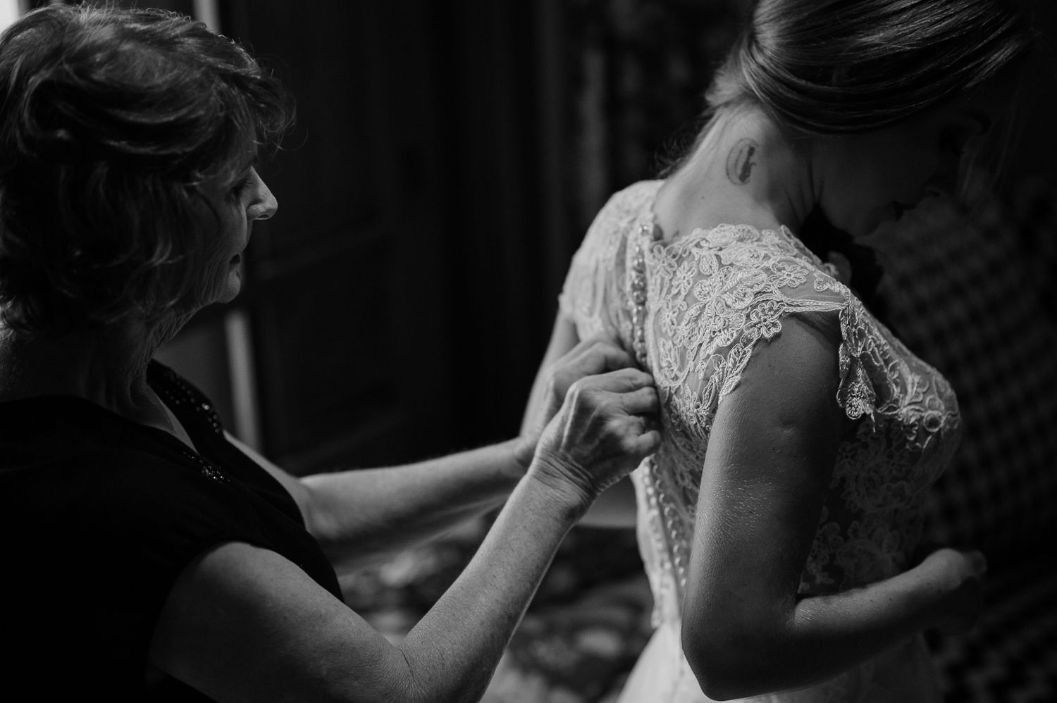 Black and white photo showing the back of a wedding dress being fastened by helping hands.