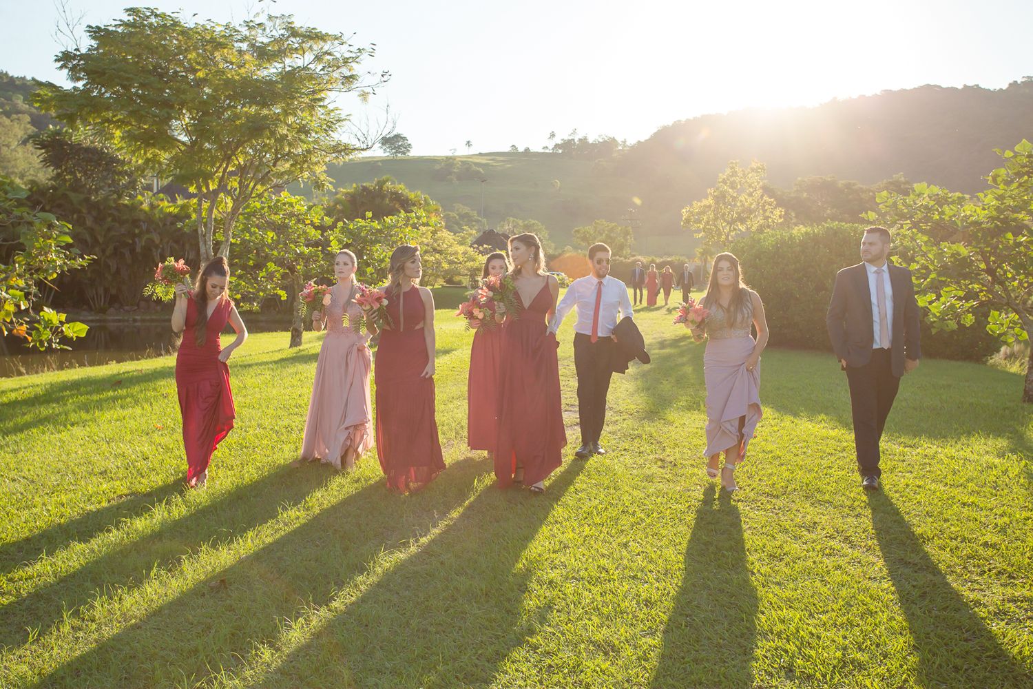 Wedding party walks together on green lawn during golden hour creating long shadows.