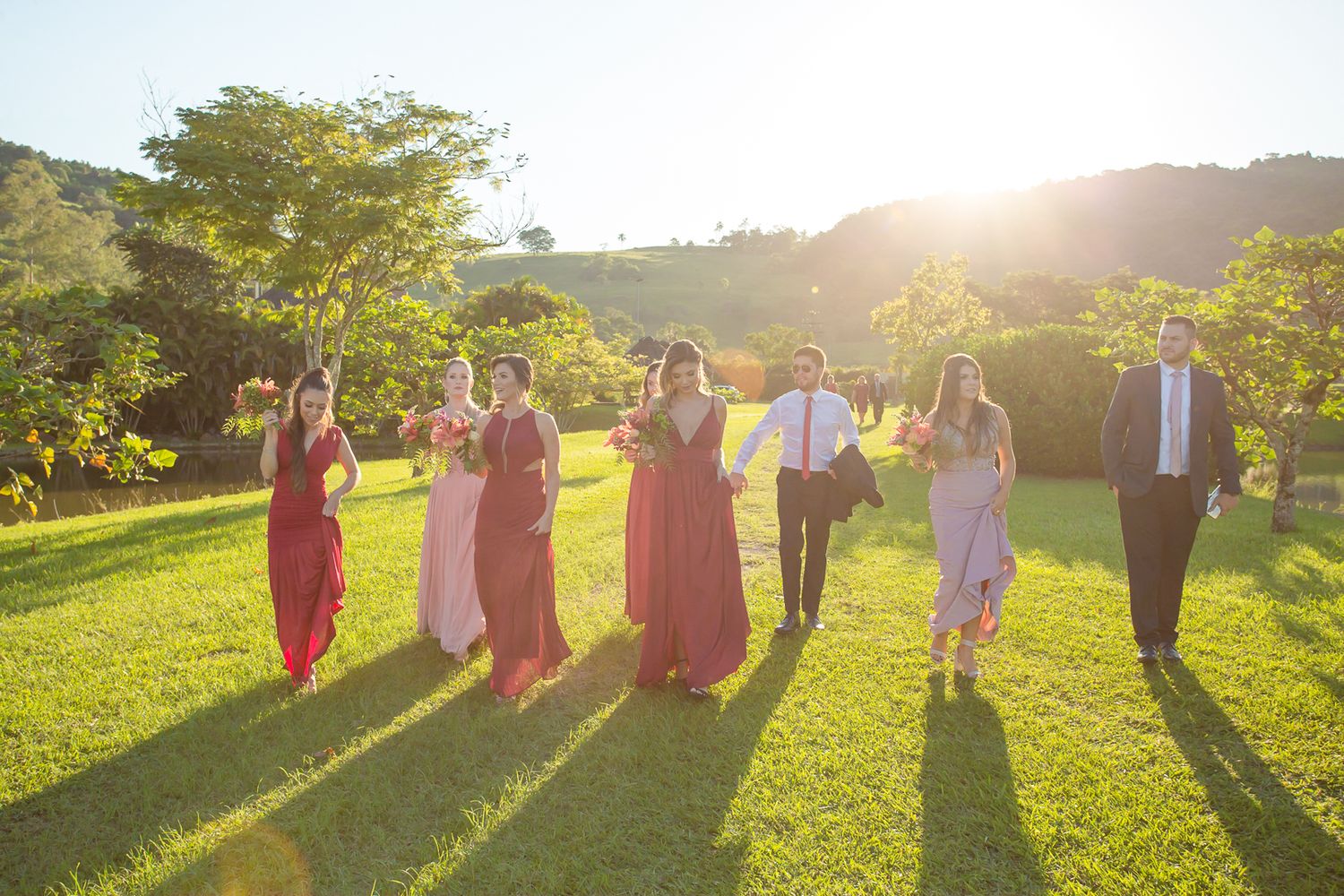 Wedding party members walk in row across sunny lawn casting long dramatic shadows.