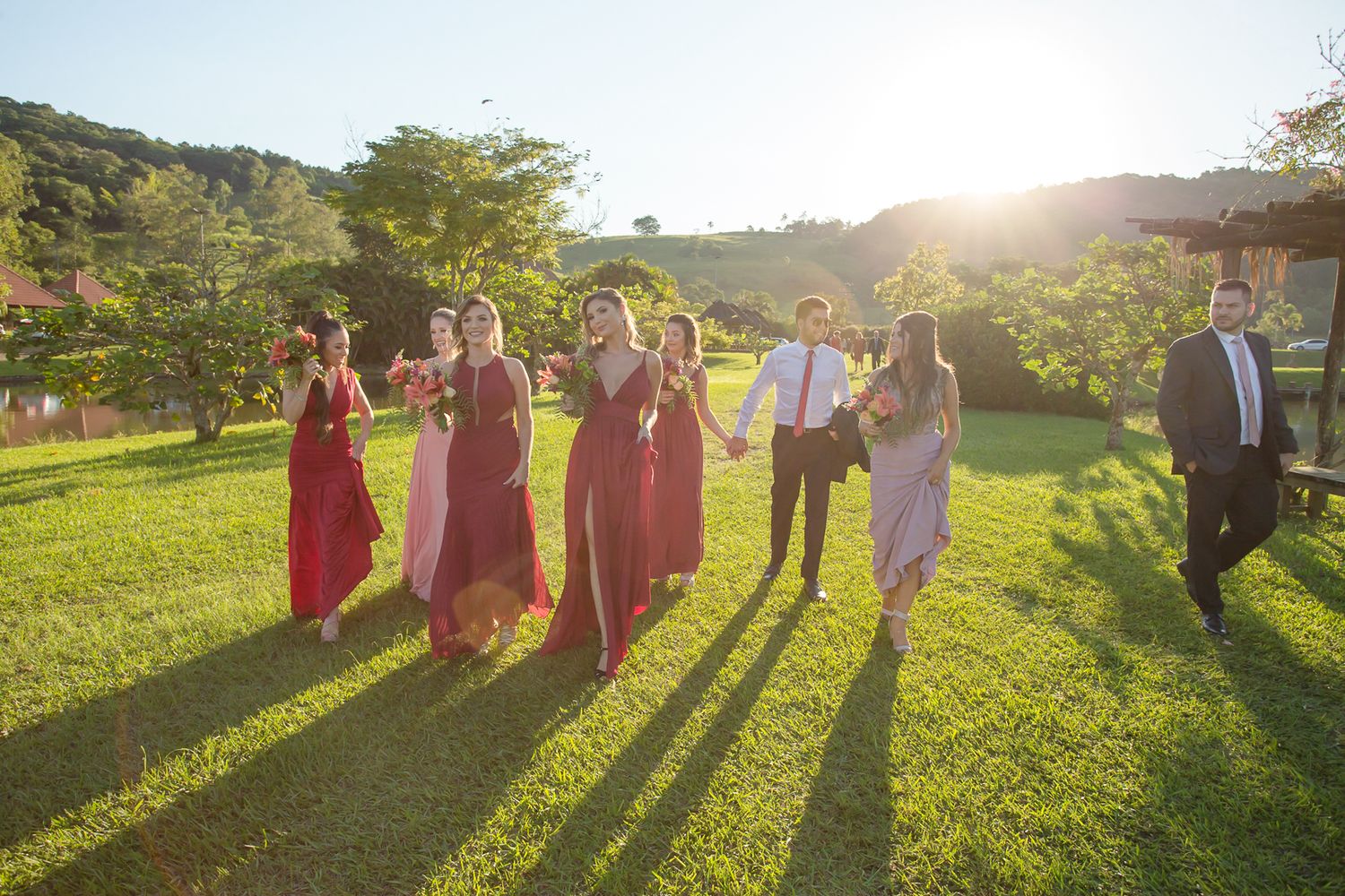 Bridal party in burgundy dresses walks across lawn during golden hour.