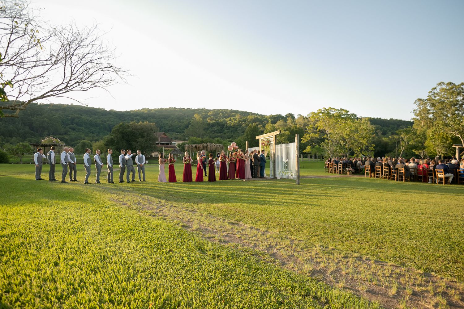 Wedding ceremony setup on green lawn with white arch and guests in formation.