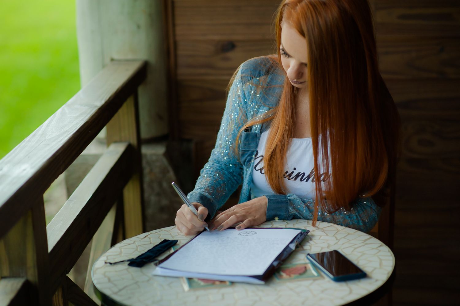 Someone writes in a notebook while sitting at a round table near a window.
