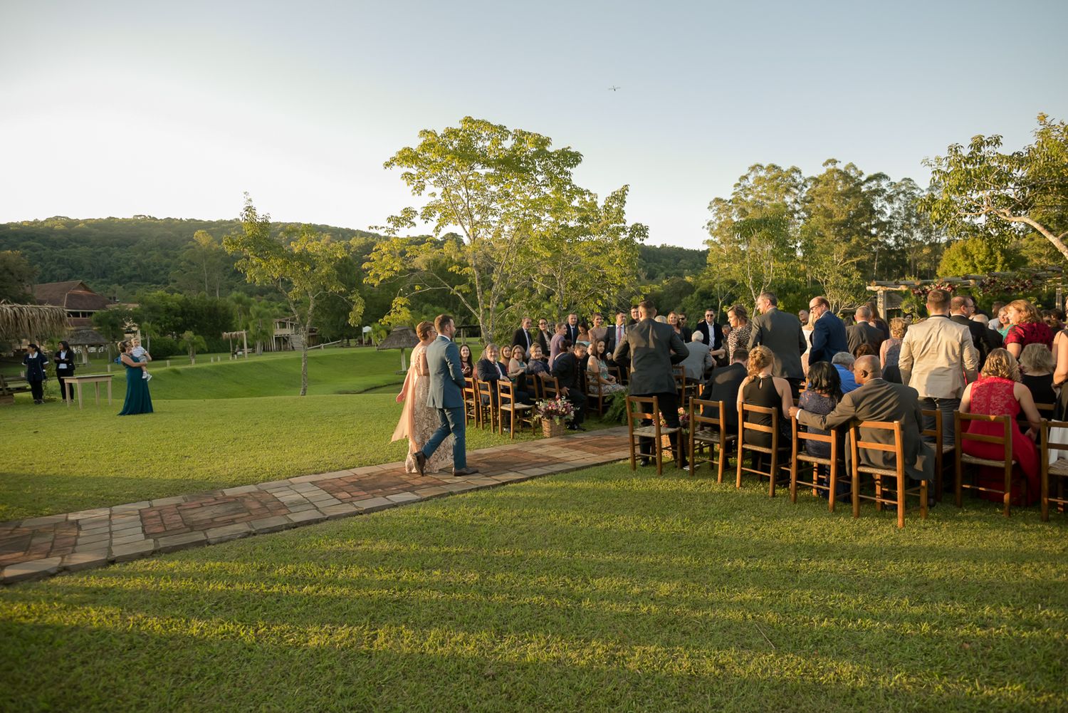 Outdoor wedding ceremony on a lush green lawn with guests seated in wooden chairs at sunset.