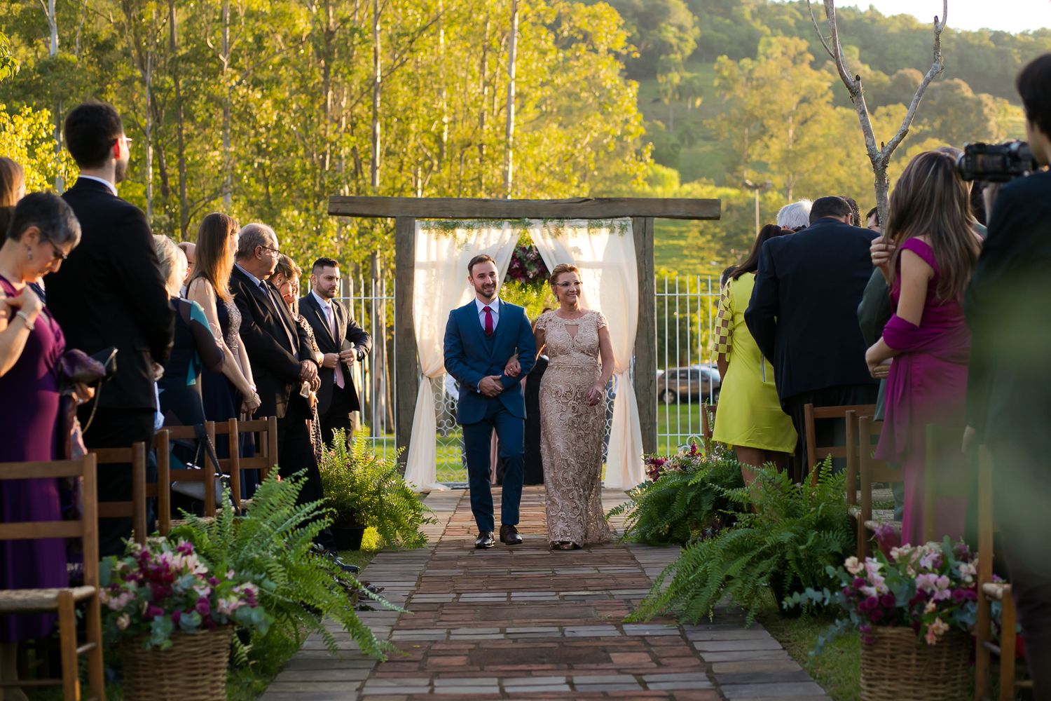 Wedding ceremony processional on stone pathway lined with potted plants and floral arrangements.