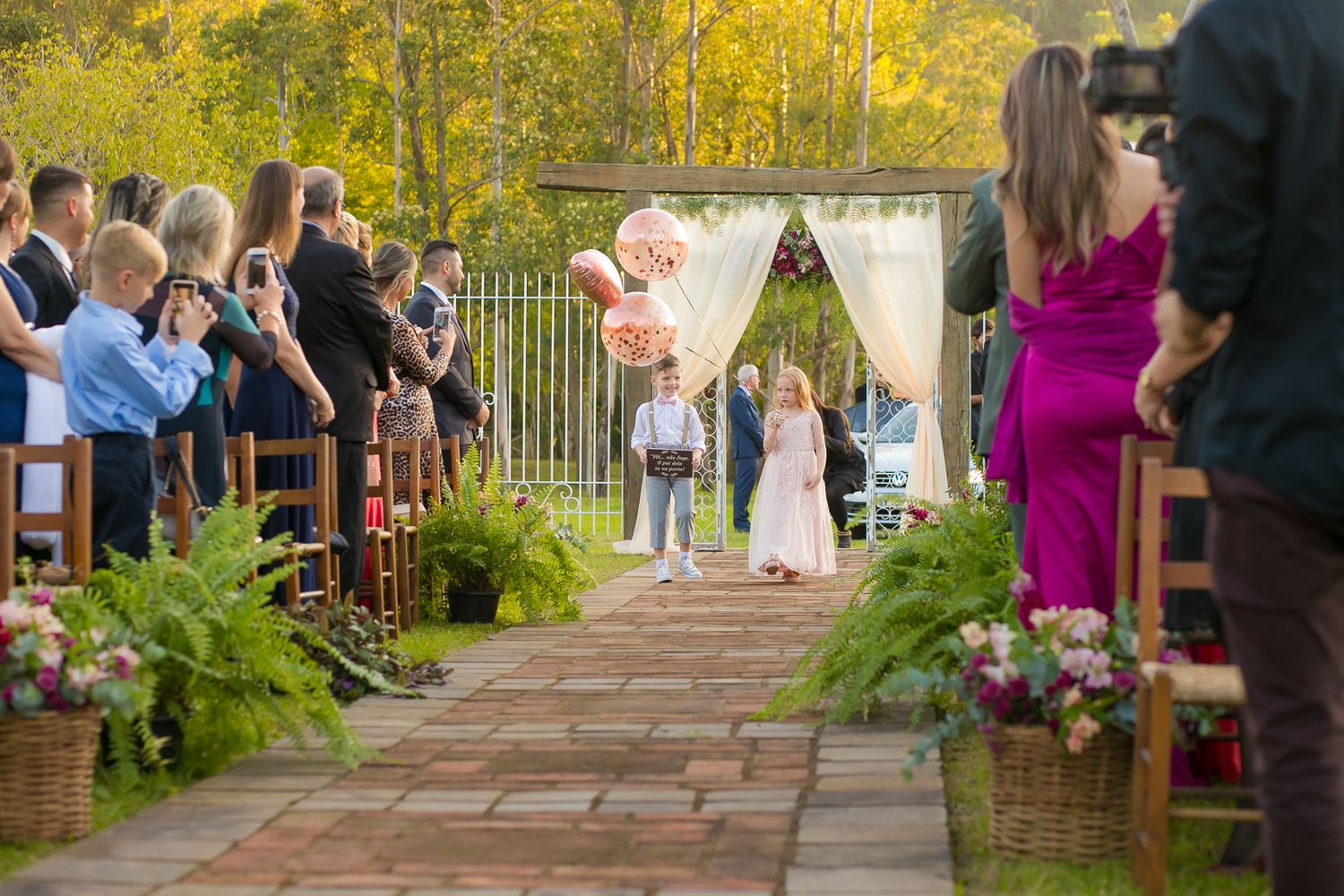 Wedding ceremony aisle lined with ferns and flowers leading to white arch backdrop.