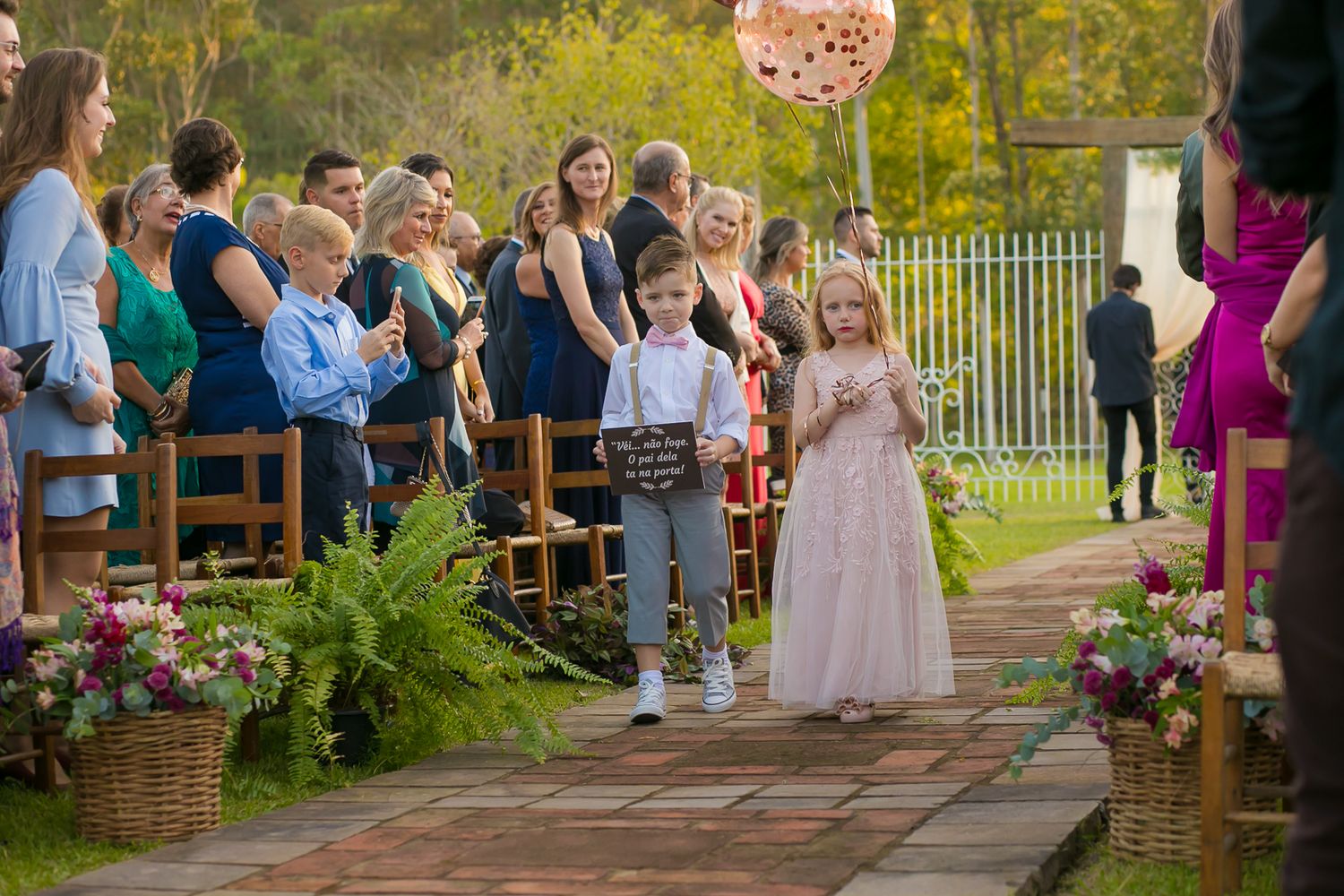 Young attendants walking down outdoor ceremony aisle lined with floral arrangements.