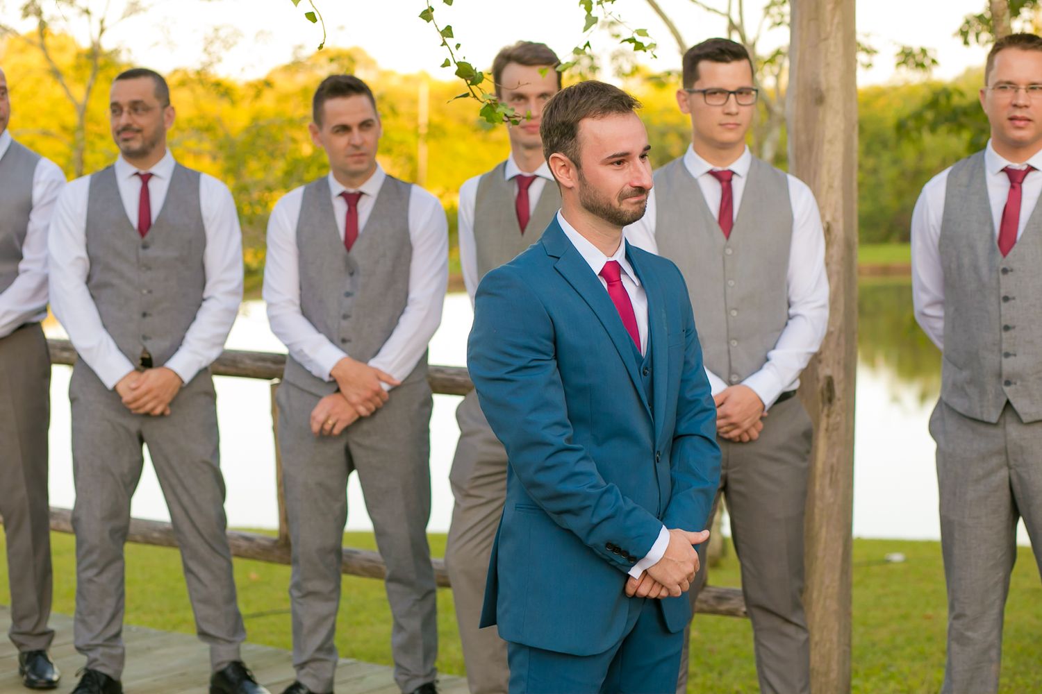 Groomsmen in grey vests and red ties standing with groom in blue suit by lakeside.