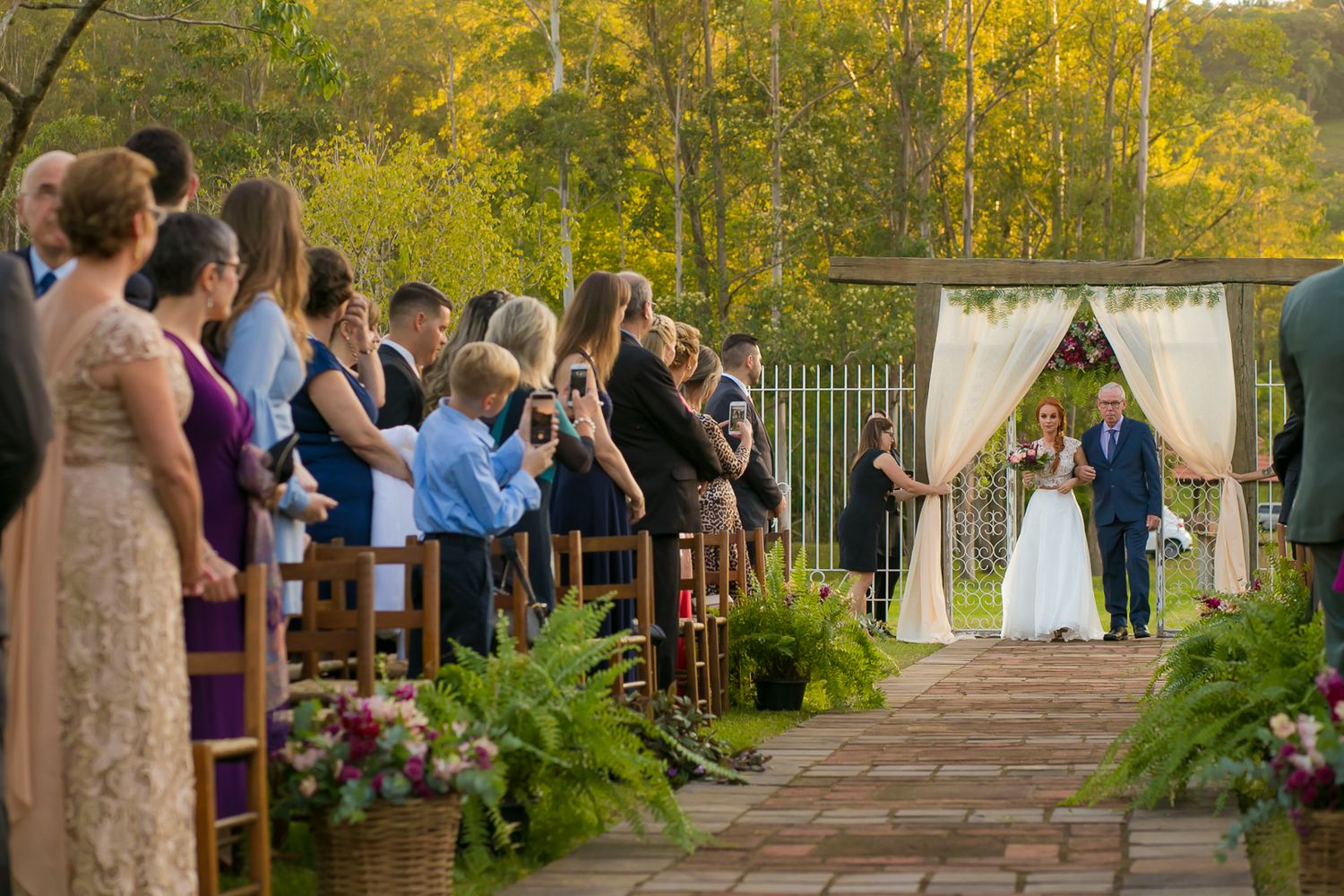 Wedding ceremony at an outdoor garden venue with guests seated along a stone pathway leading to a white draped arch.