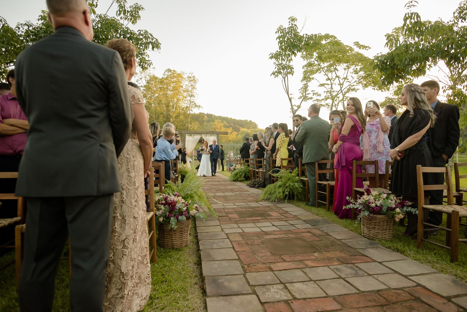 Outdoor wedding ceremony with guests seated along stone path surrounded by lush greenery and trees.