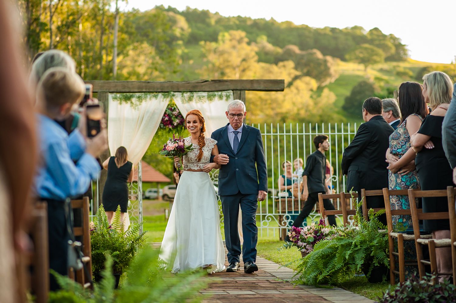 Evening outdoor wedding ceremony with golden sunlight filtering through trees and white ceremony arch.