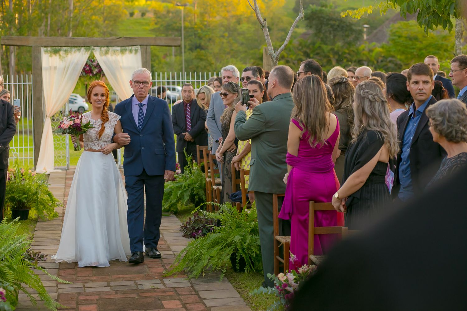 Guests look on during a traditional wedding processional on stone pathway surrounded by ferns.