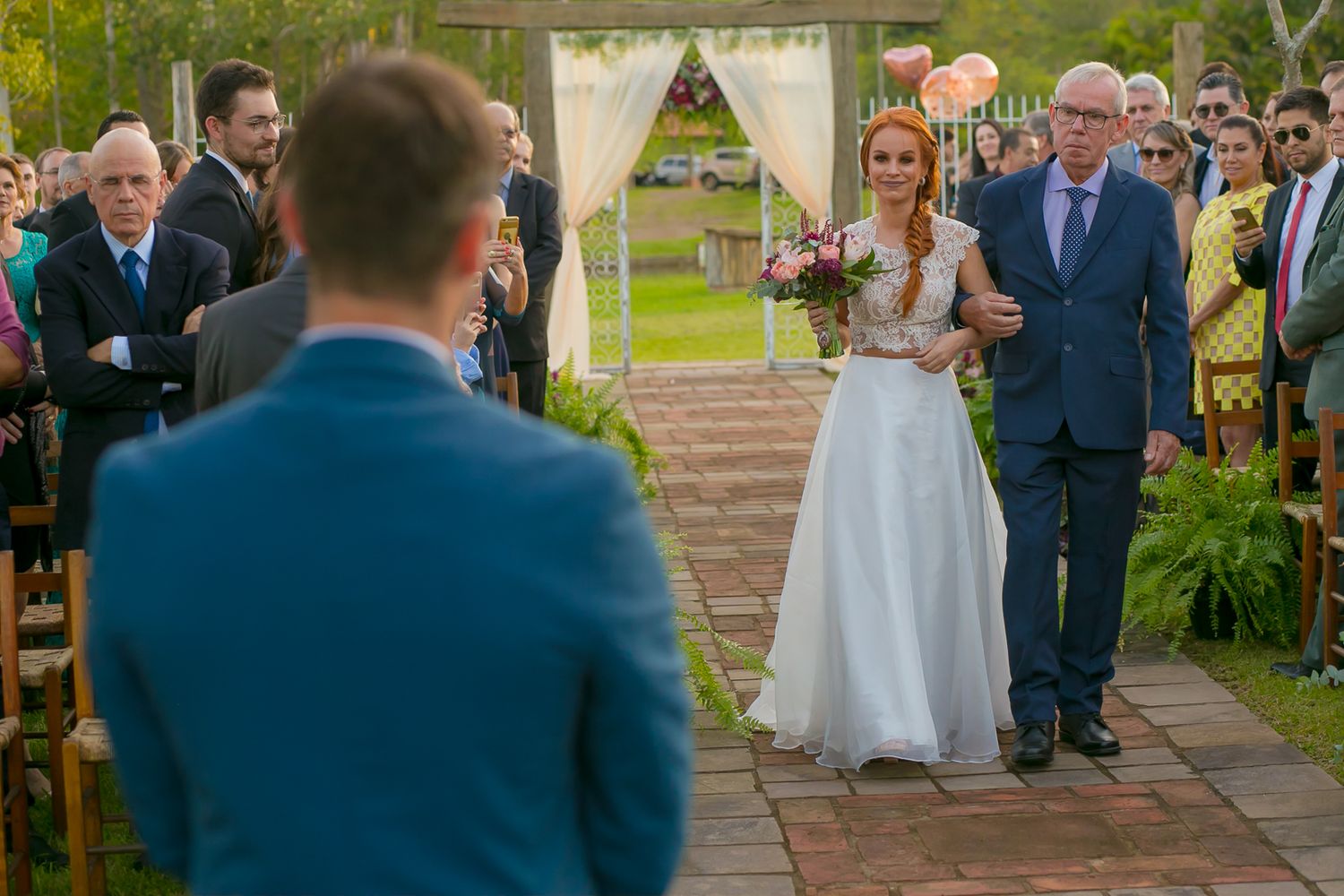 Wedding processional on stone pathway with white draped arch and natural garden backdrop.