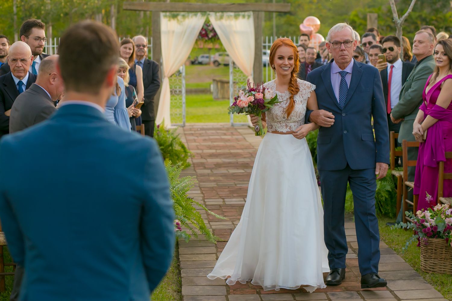 Outdoor garden wedding ceremony with stone pathway and white draped arch against green landscape.