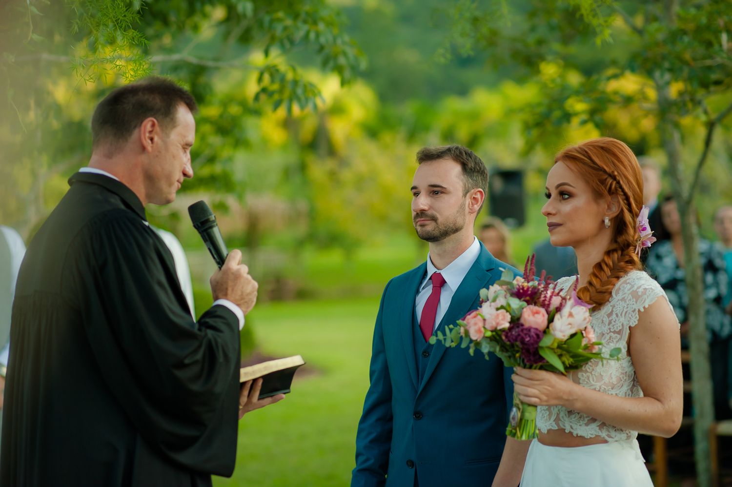 Outdoor wedding ceremony with officiant speaking to couple holding bouquet in garden setting.