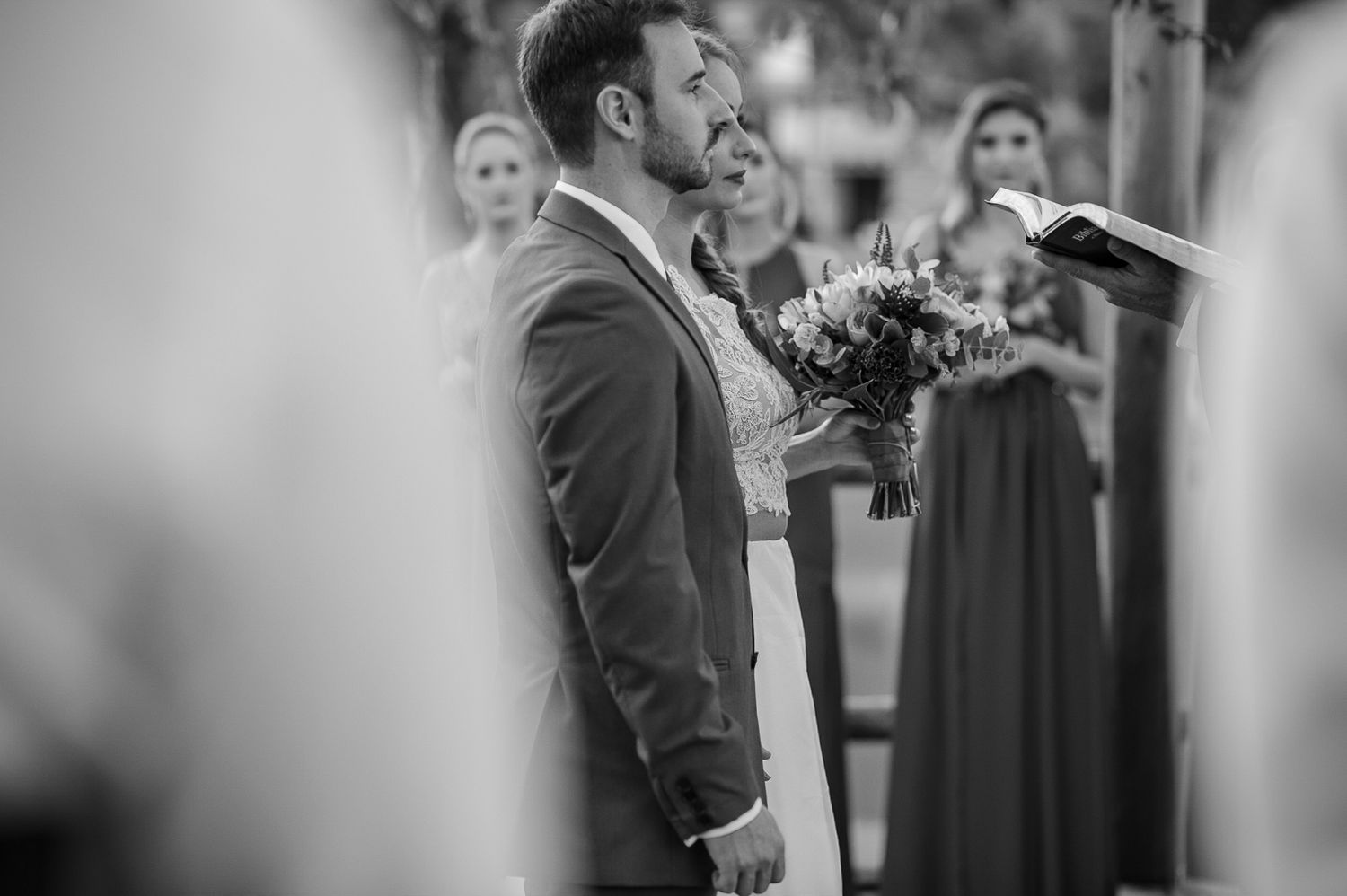 Black and white image of a wedding ceremony moment with guests and floral bouquet in the background.
