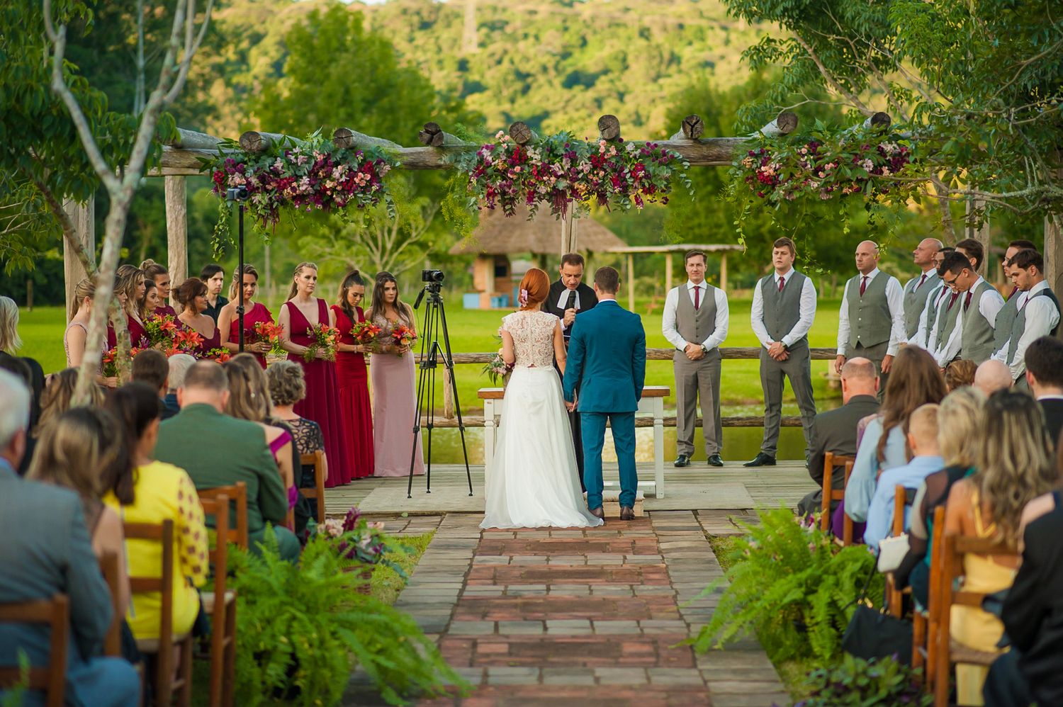 Beautiful outdoor wedding ceremony with floral arch, red bridesmaid dresses, and seated guests.