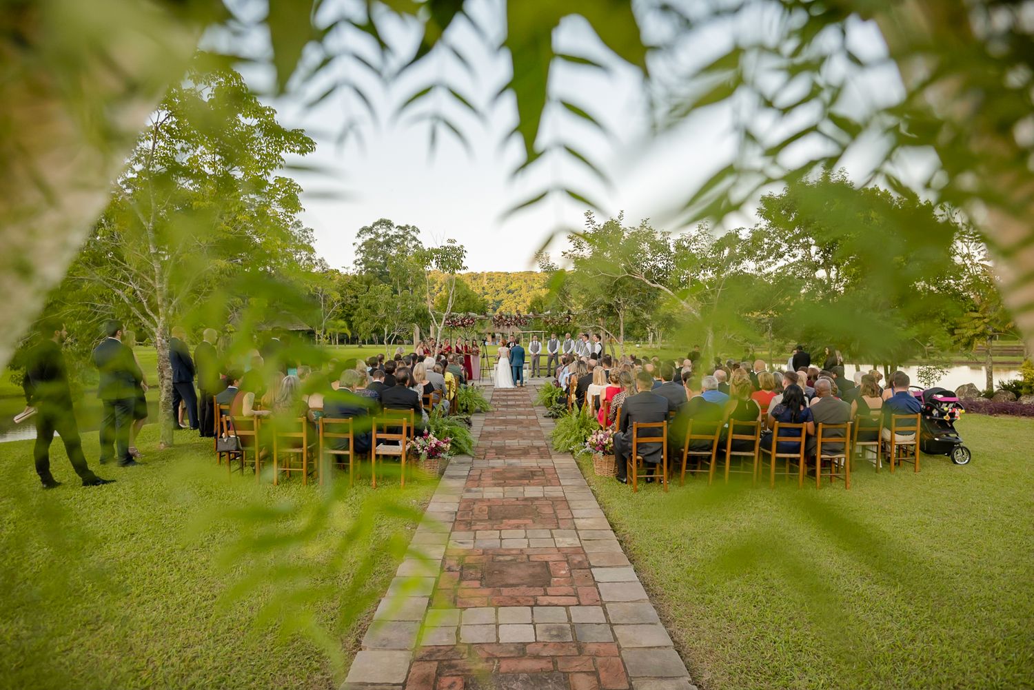 Scenic garden wedding ceremony view through leaves with brick pathway and seated guests.