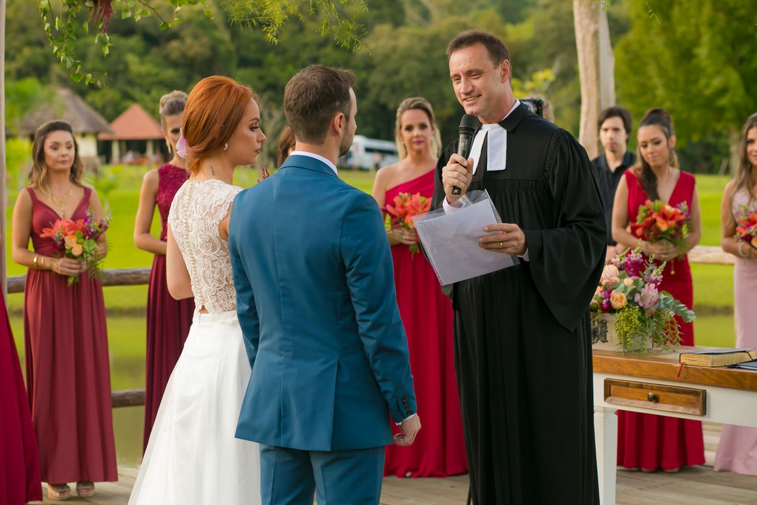 Wedding ceremony in progress with officiant in black robes and bridesmaids in burgundy dresses.