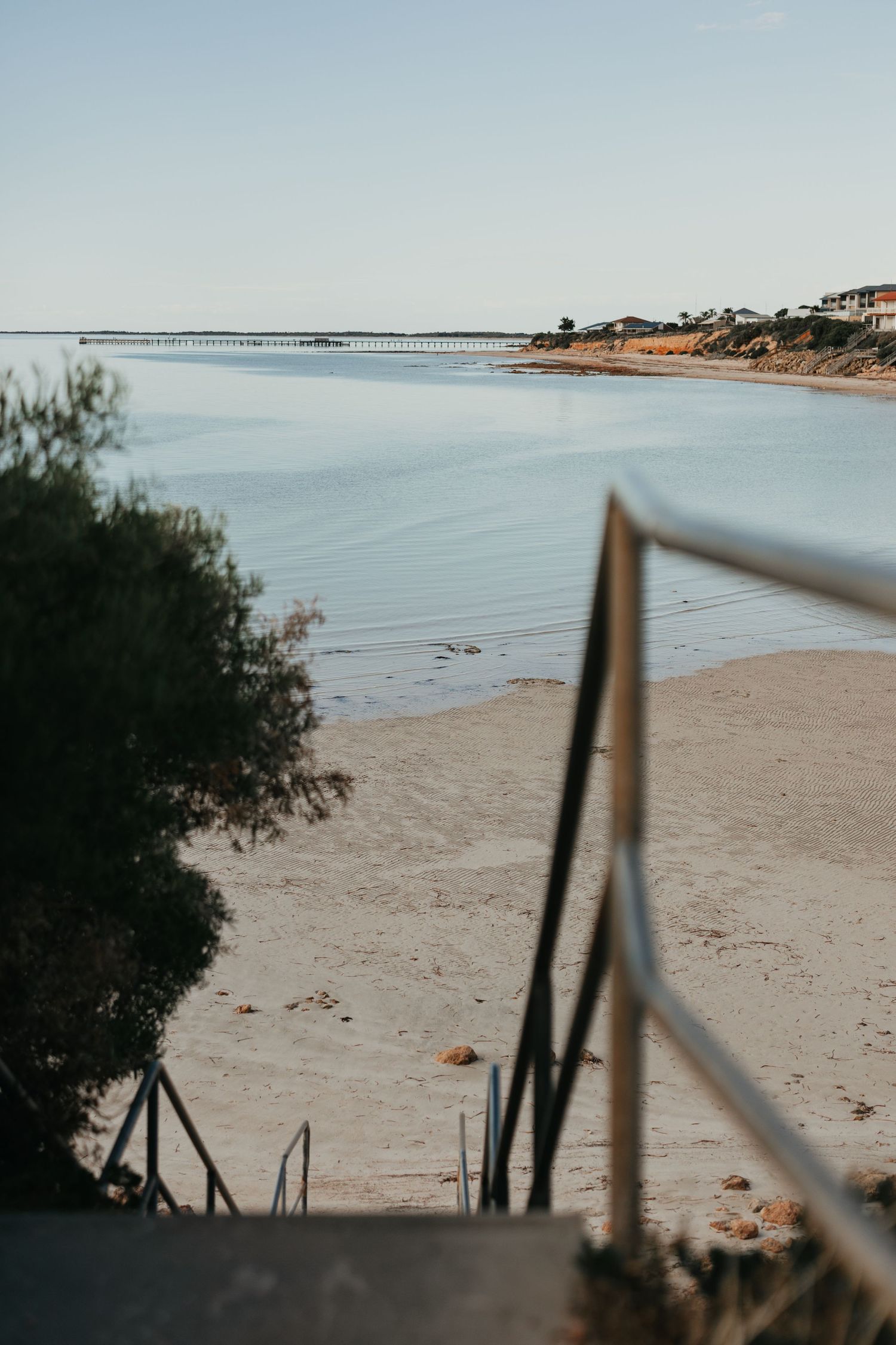 View of a long pier stretching into calm blue waters from beach steps.