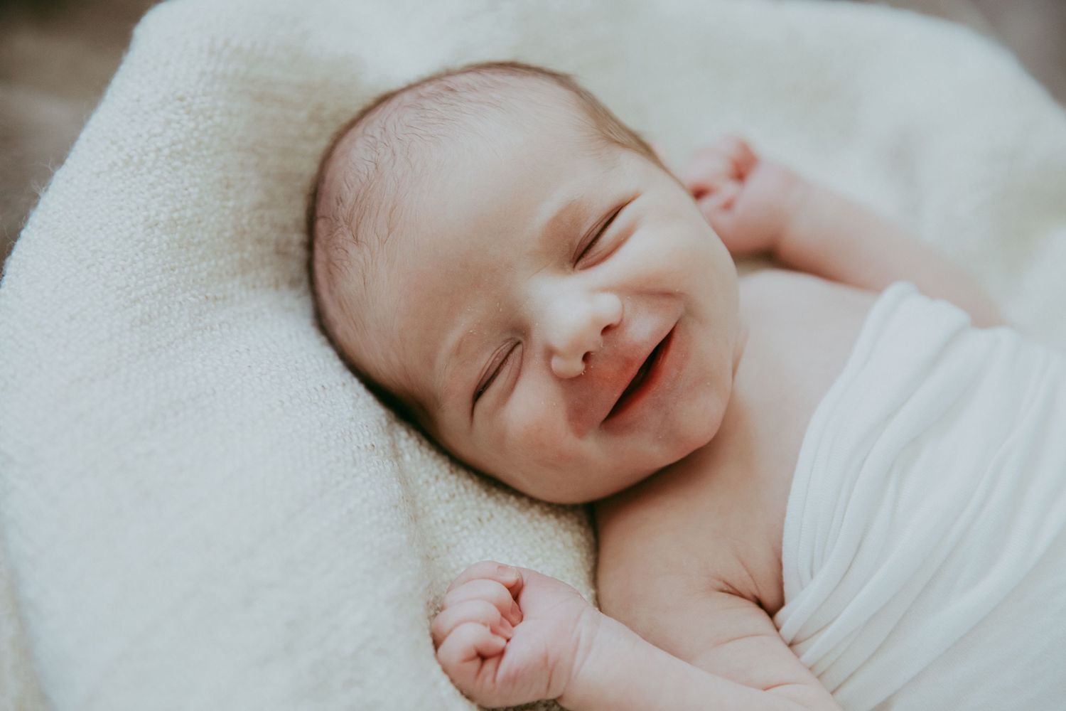 Peaceful sleeping newborn wrapped in white swaddle smiles contentedly.