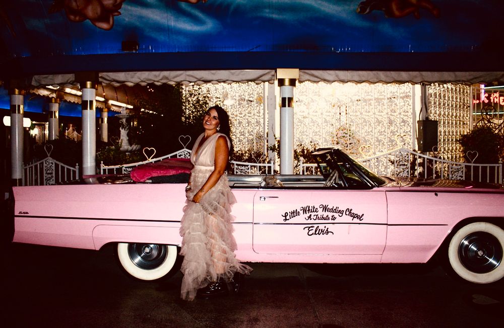 Woman in elegant gown posing beside a pink Elvis Presley Cadillac convertible at a Las Vegas attraction.