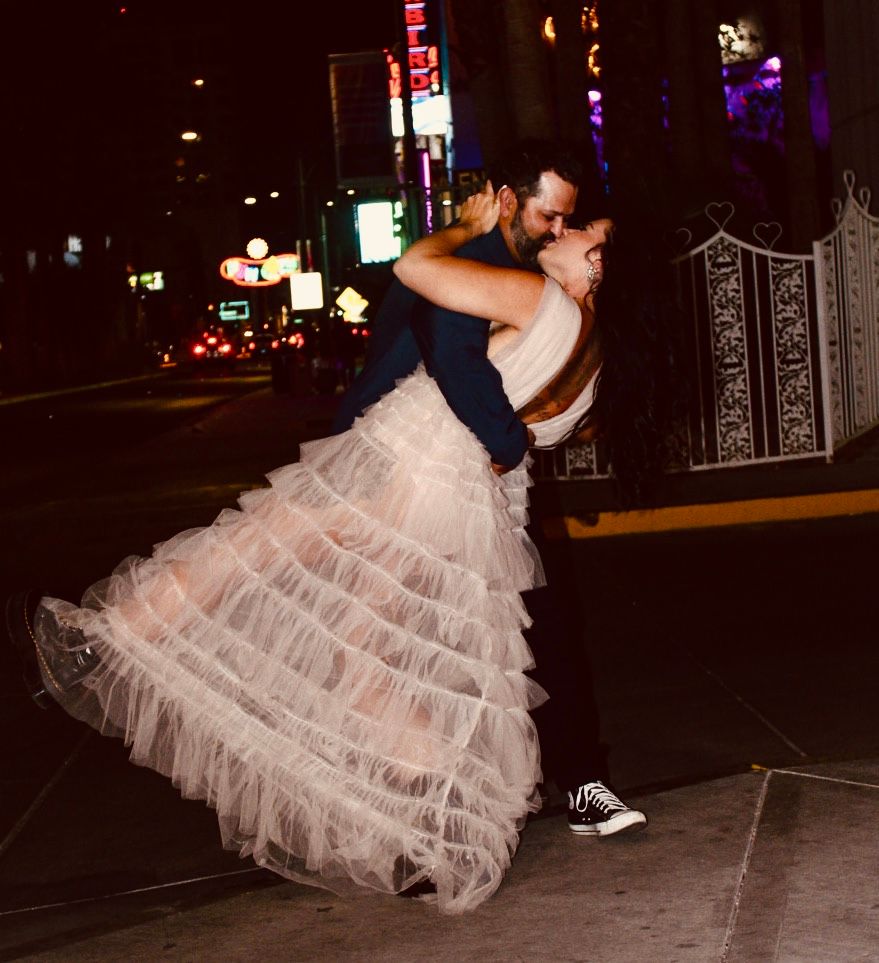 A groom lifts his bride in a ruffled tulle skirt on a city street at night, surrounded by glowing neon lights.