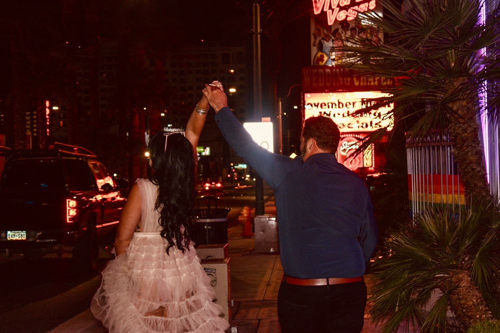 Couple in wedding attire holding hands while walking along a neon-lit Las Vegas Strip at night.
