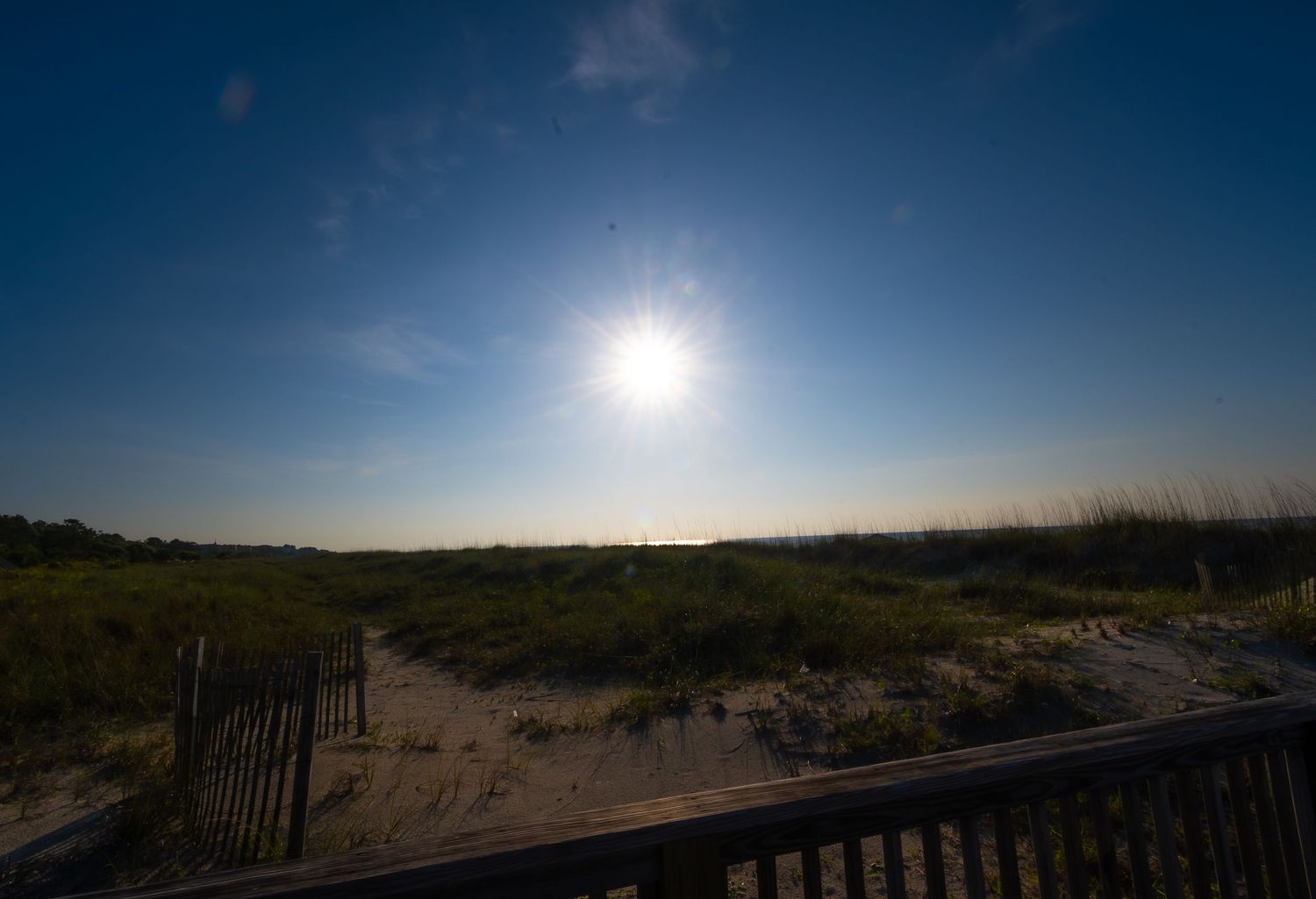 Bright sun illuminates a beach boardwalk with sand dunes under a deep blue sky.