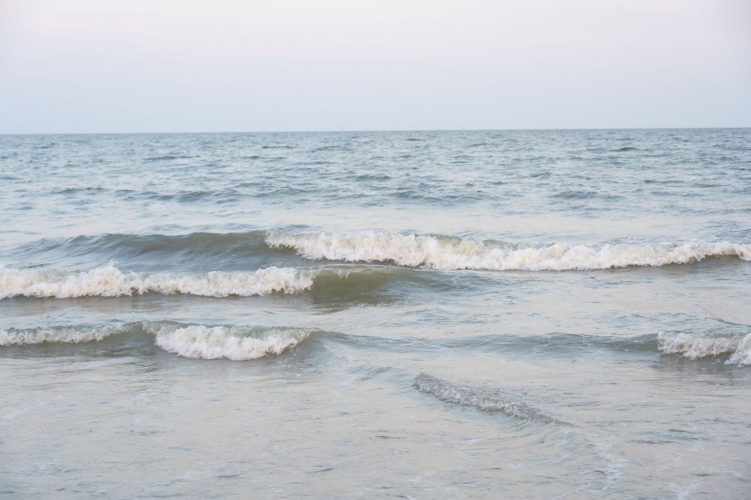 Rolling waves approach shoreline under overcast sky in muted tones.