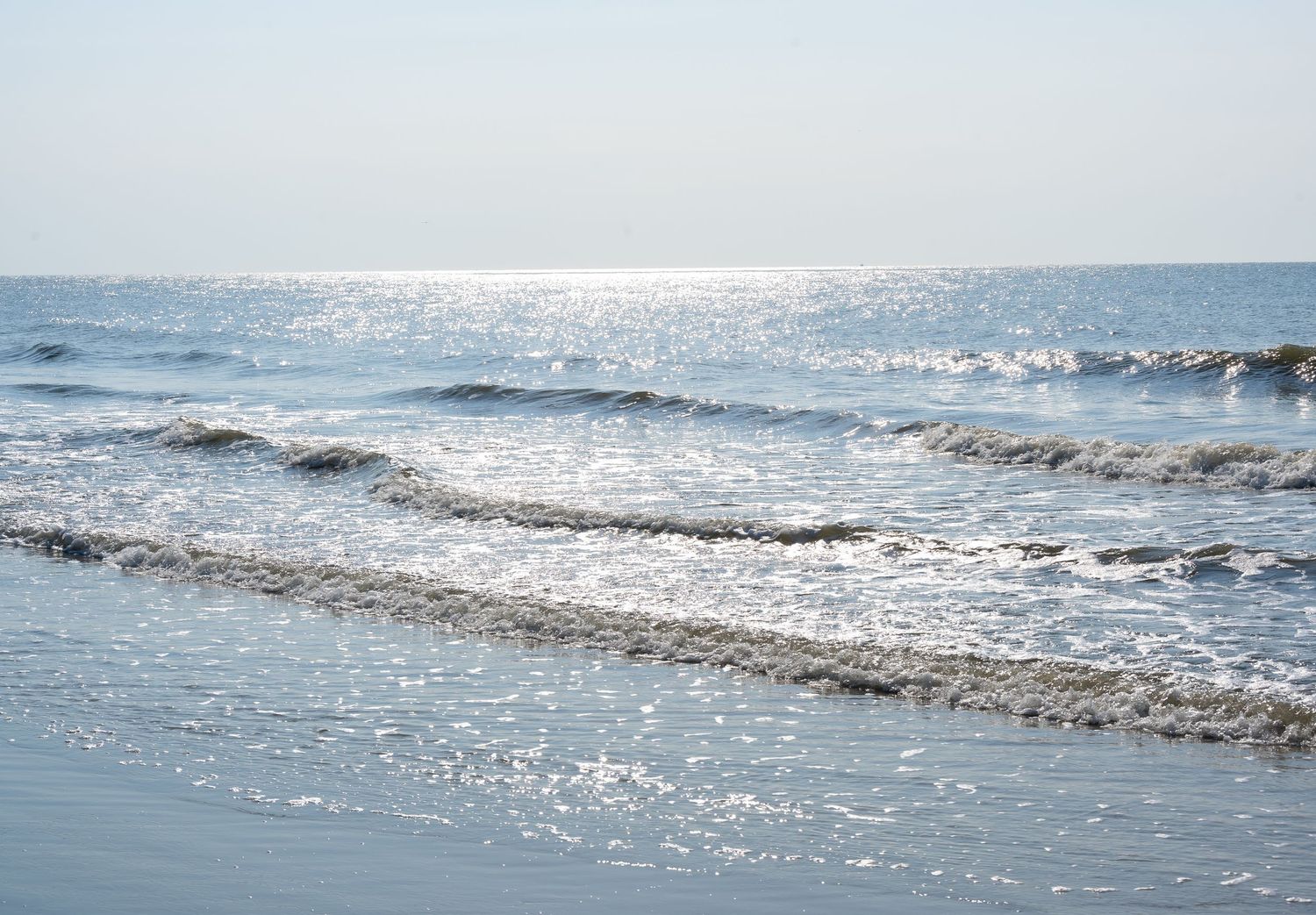 Sunlight sparkles on rolling ocean waves approaching a sandy beach.