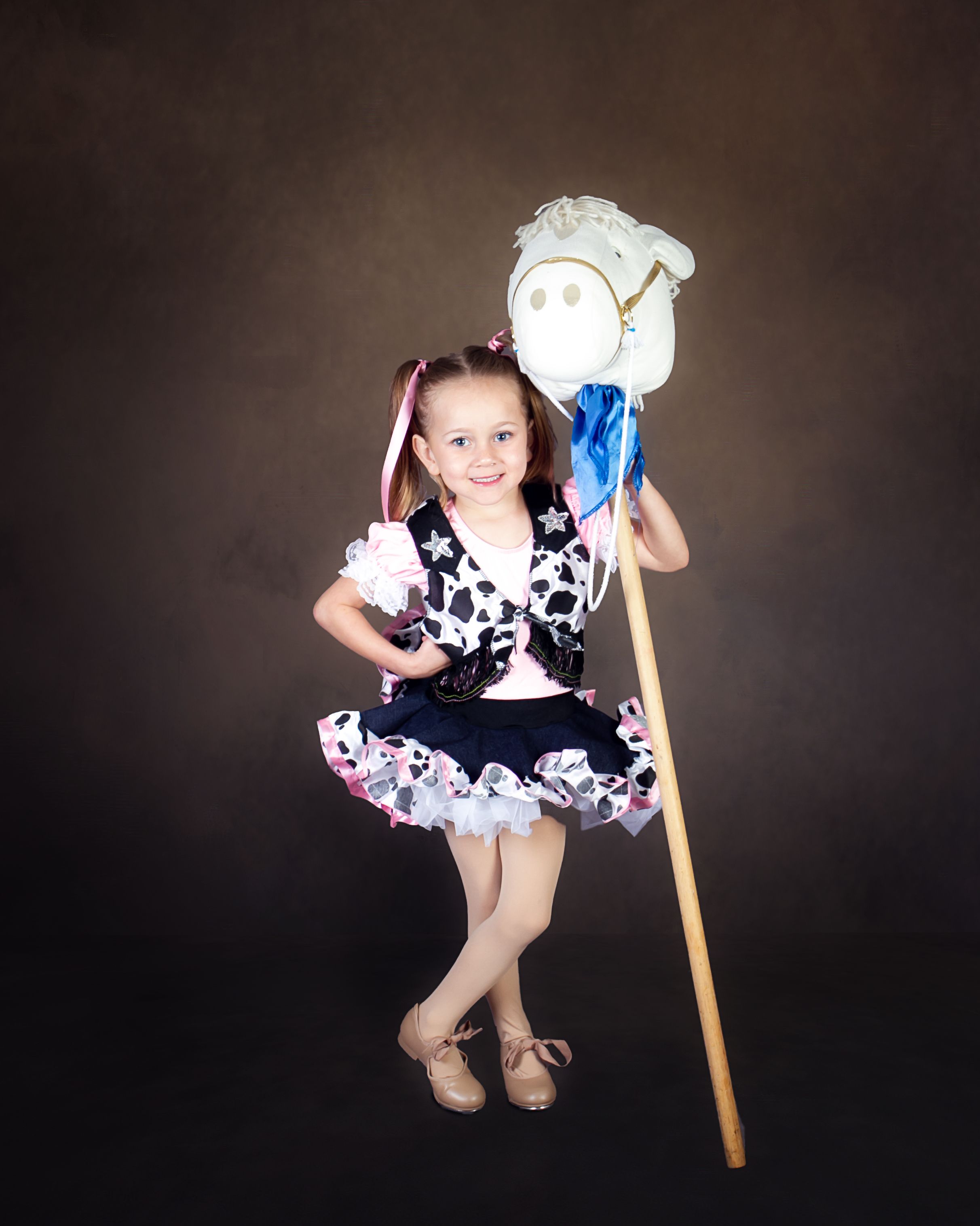 A young dancer in a black and white polka dot dress with tulle petticoat poses with a hobby horse stick prop against dark background.