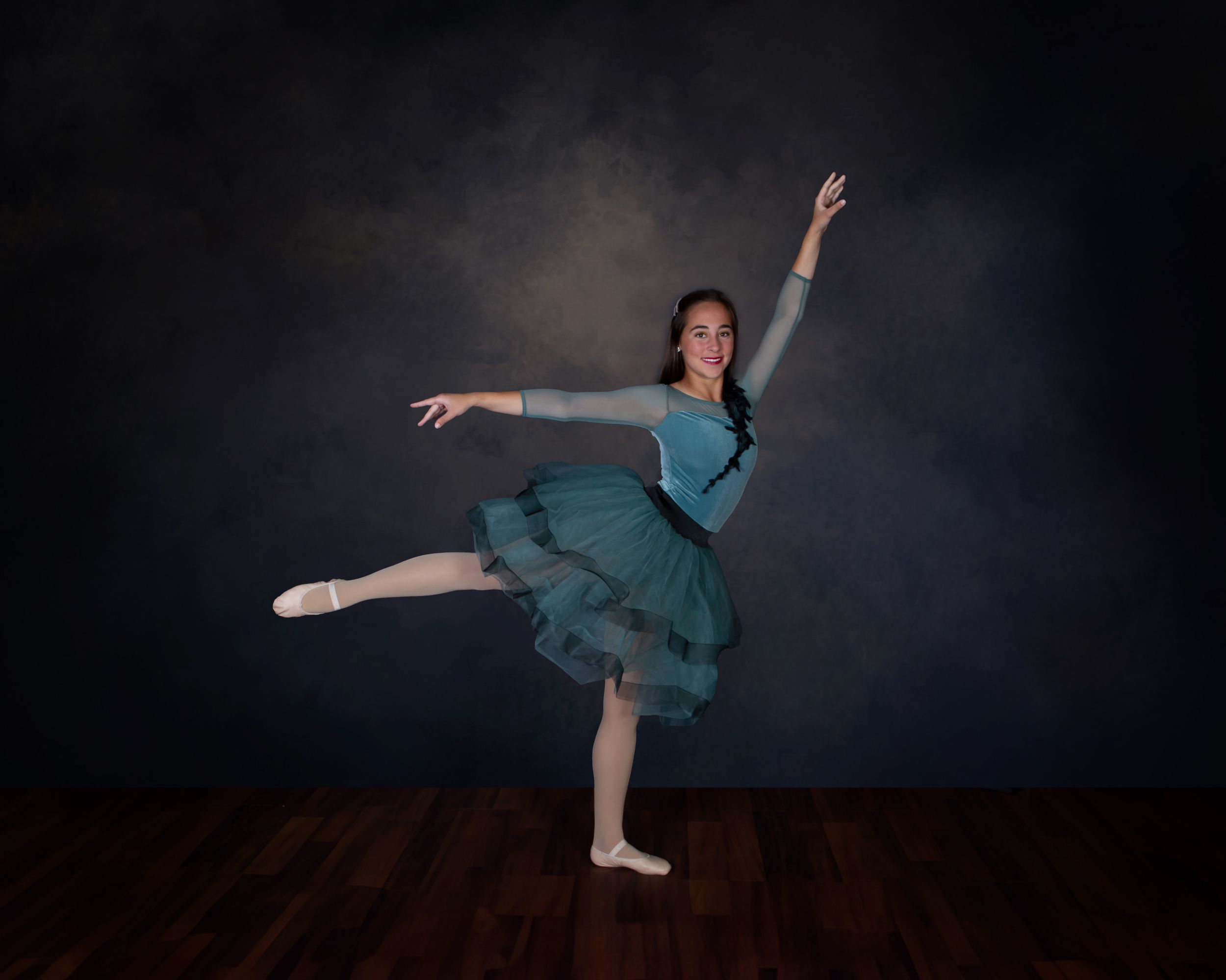 Ballet dancer in teal dress performs graceful pose on dark wooden stage.
