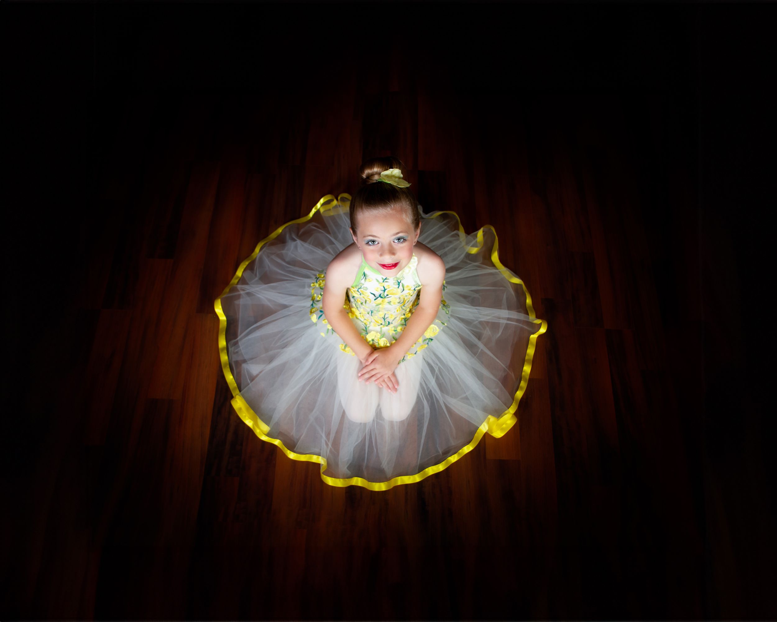 Ballet dancer in illuminated white tutu photographed from above on dark wooden floor.