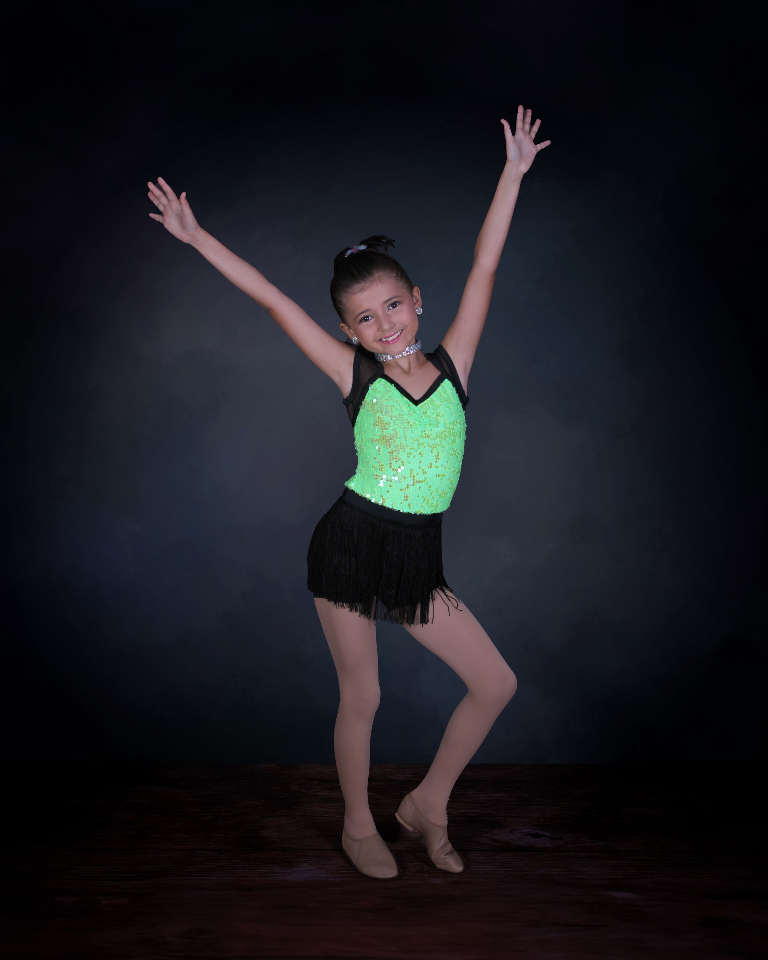 A dancer in a sparkly green leotard and black fringe skirt strikes an energetic pose against a dark background.