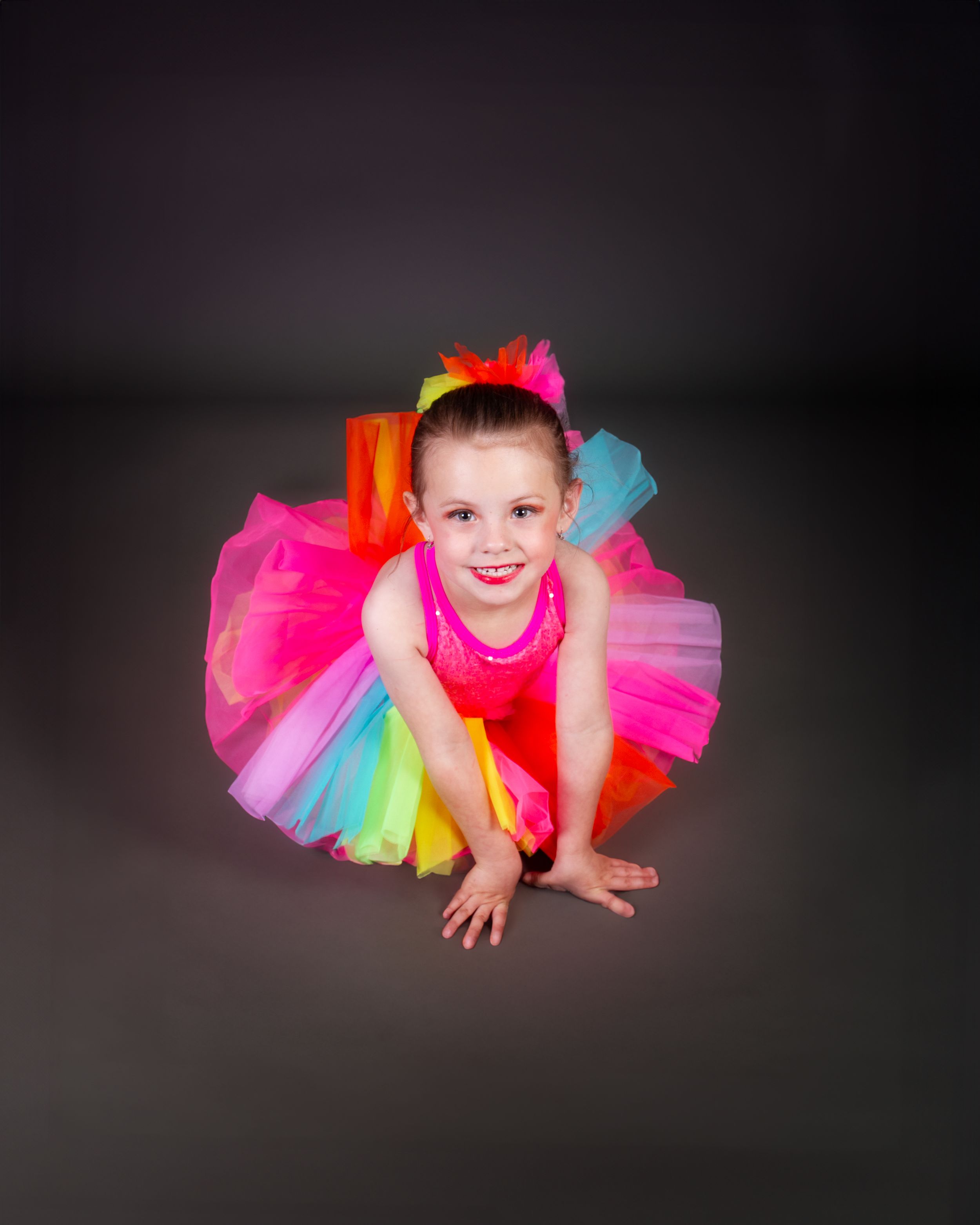 A young dance student wearing a colorful rainbow tutu dress poses on a dark backdrop during a studio photoshoot.