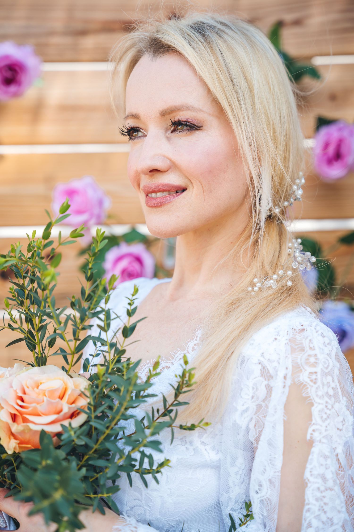 Elegant bride in white lace dress poses with roses against wooden backdrop.
