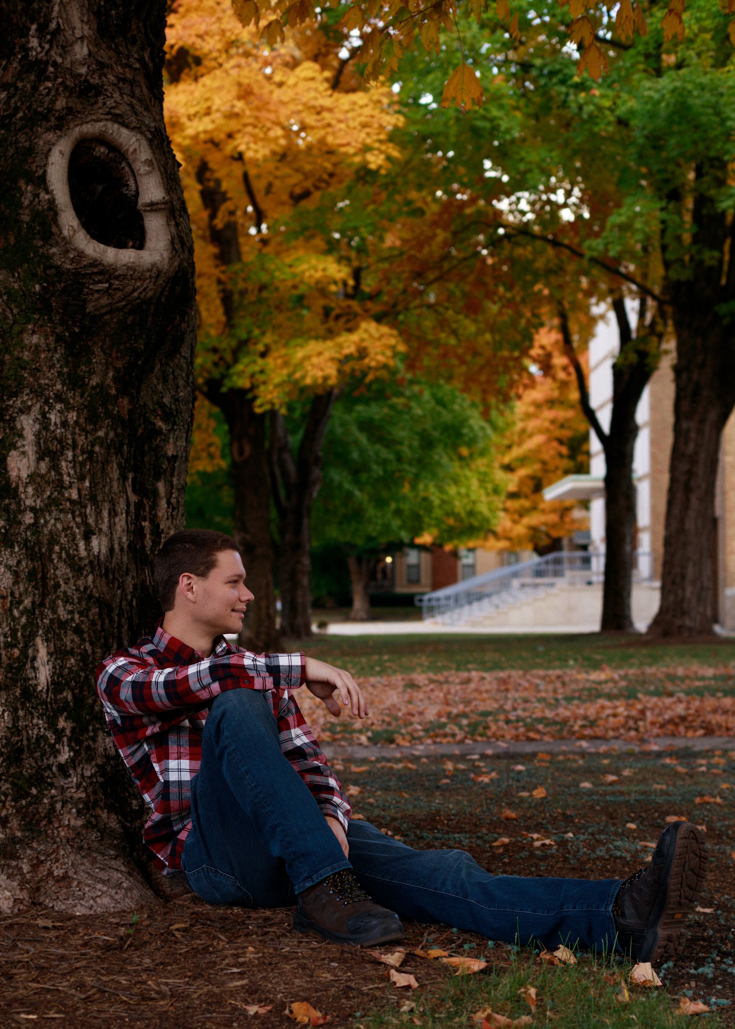 Person in plaid shirt and overalls lounging against tree trunk during autumn with fallen leaves on ground.