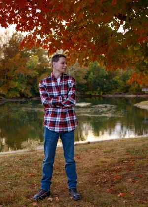Person in plaid shirt and jeans poses near a pond with autumn foliage in the background.