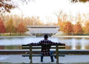 Wide shot of person sitting alone on park bench by lake during fall season.