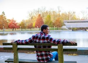 Person relaxing on park bench by lake with autumn trees in background during sunset.