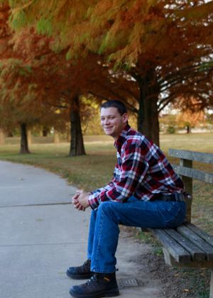 Person in plaid shirt and jeans sitting on bench surrounded by autumn trees.