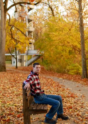 Person in plaid shirt sitting on wooden bench surrounded by fallen autumn leaves.