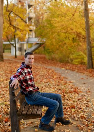 Person in plaid flannel and jeans sitting casually on wooden bench amid fall leaves.