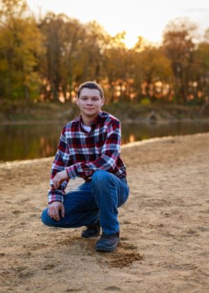 Person sitting on sandy beach near lake during autumn sunset with trees in background.