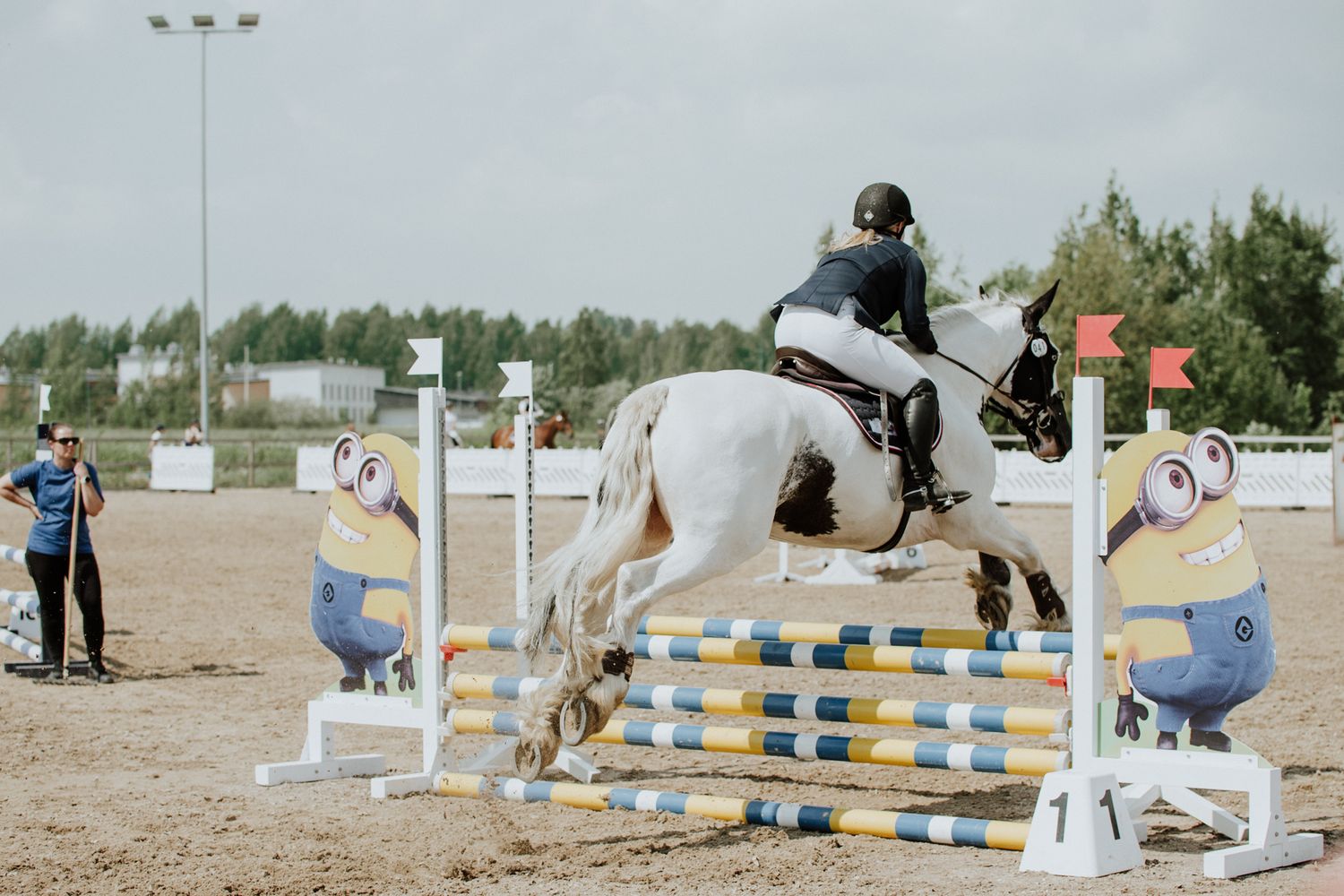 Equestrian jumping over white obstacles in competition arena with Minion decorations.