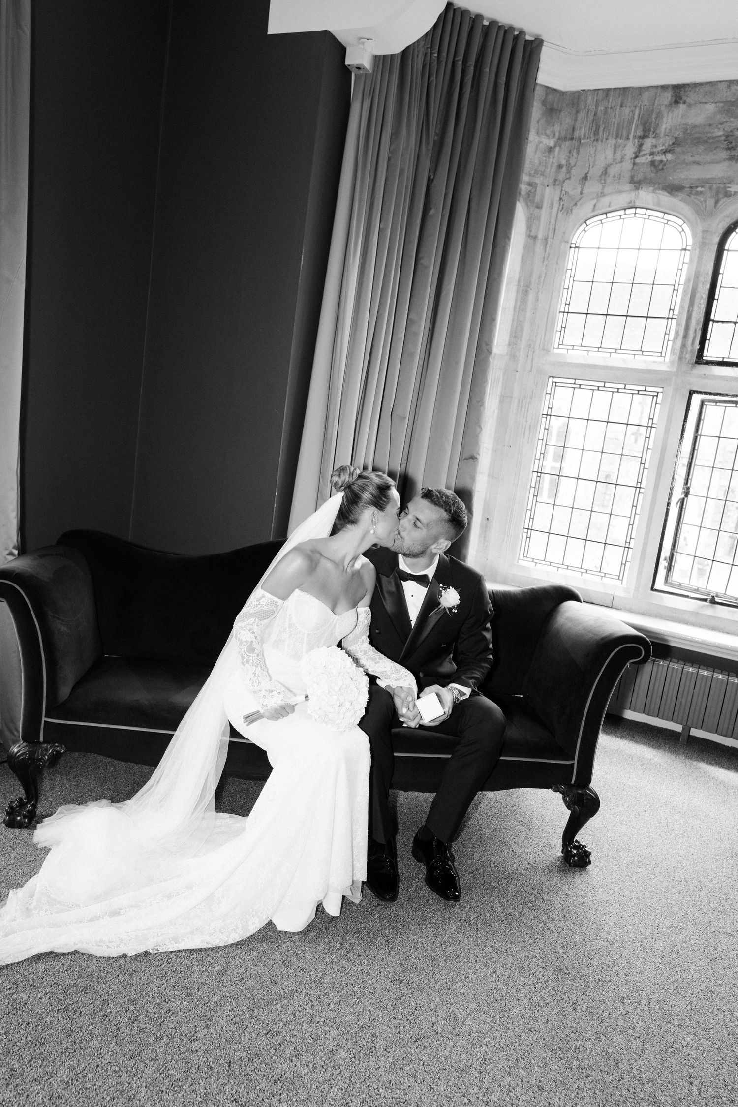 Black and white photo of a bride and groom sharing a kiss while seated on an elegant sofa near arched windows.