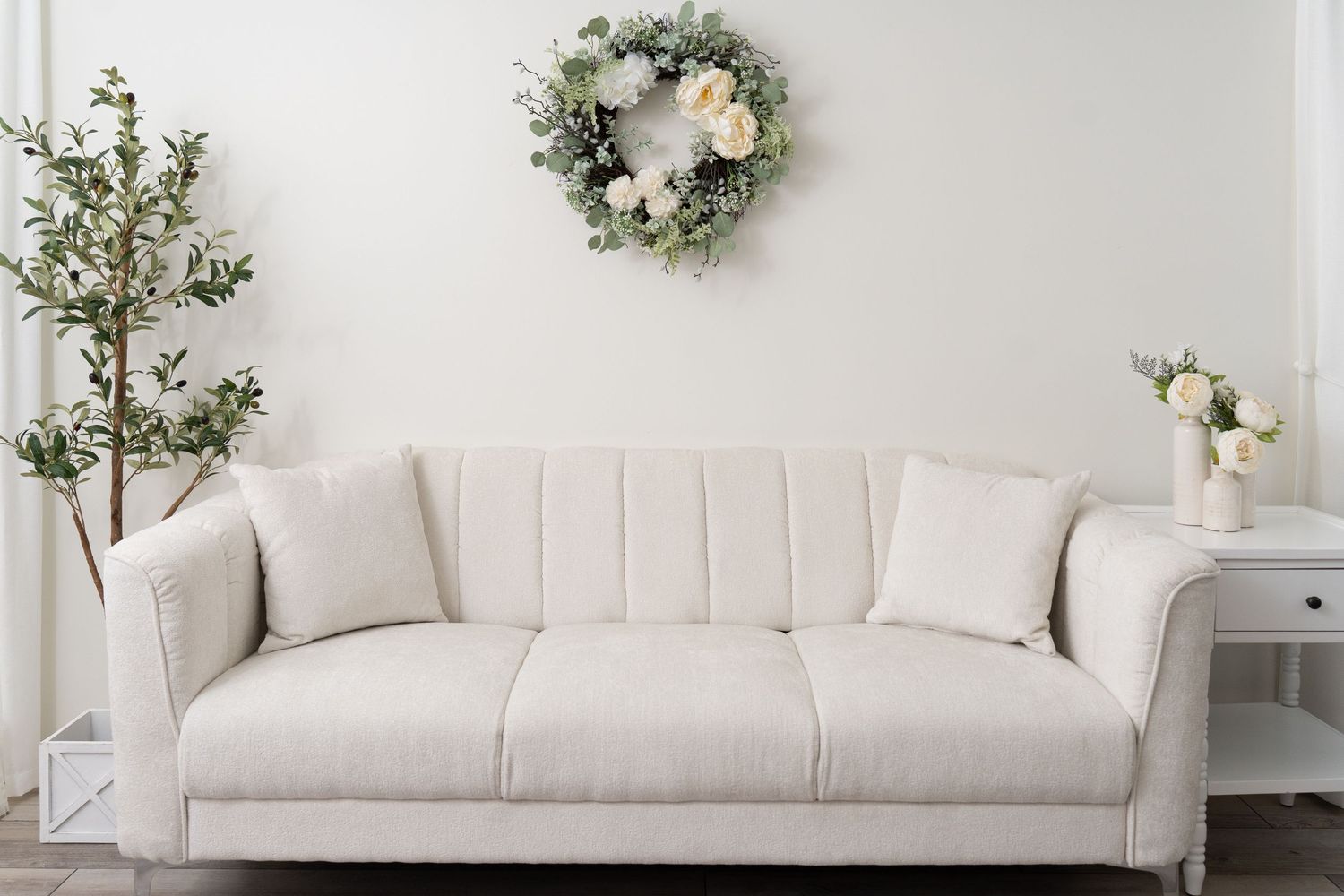 Bright living room with a cream tufted sofa, eucalyptus wreath, and floral arrangements on white walls.